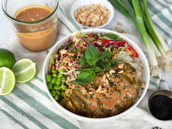 White ceramic bowl holding a chicken meal with thai seasoning, surrounded by limes, green onions, soy sauce, and ground peanuts.